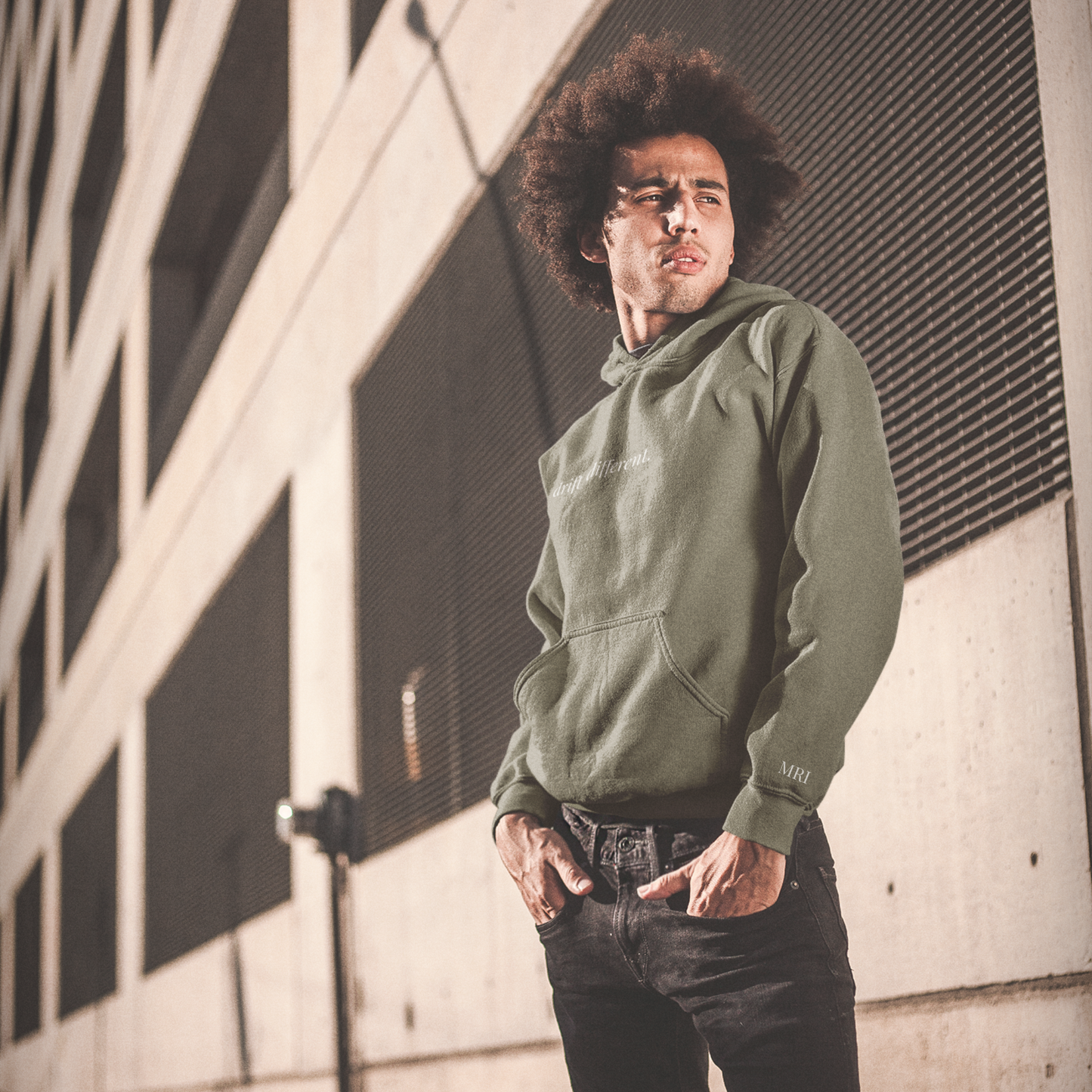 Man wearing a military green drift different. hoodie, eyes squinted by the sun, standing against a building wall.