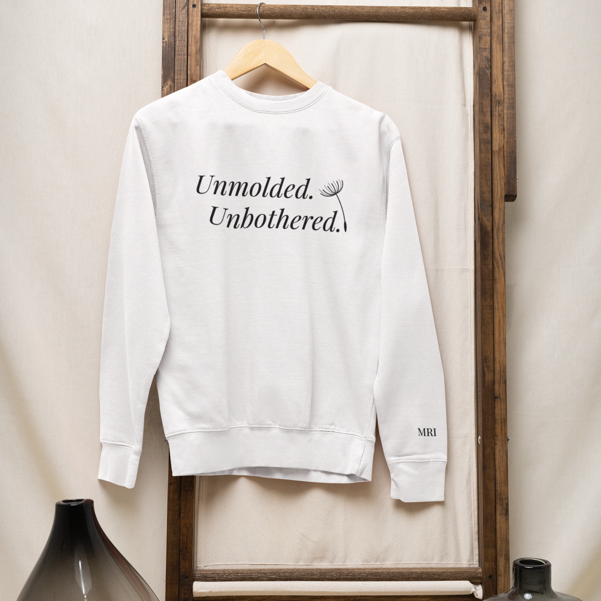 Front view of Unmolded. Unbothered. classic sweatshirt in white with bold text design on a natural color wooden hanger on a dark wooden ladder.