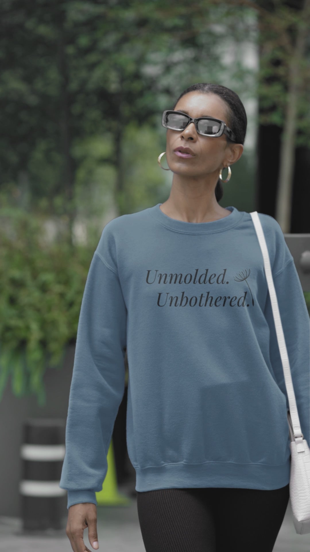Video of a woman wearing an indigo blue Unmolded. Unbothered. sweatshirt, standing in a park.